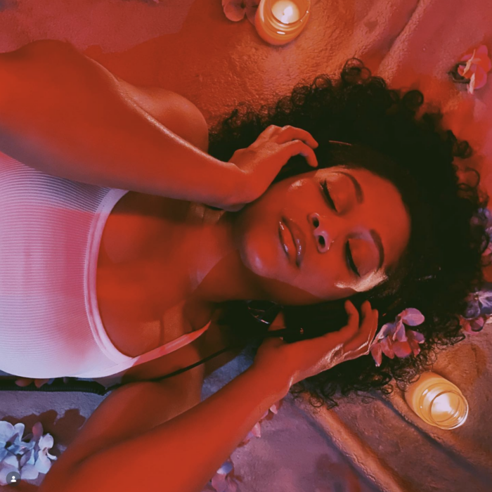 R&B artist Whitney J lying down in warm red lighting, wearing headphones and a white tank top surrounded by candles and flowers.